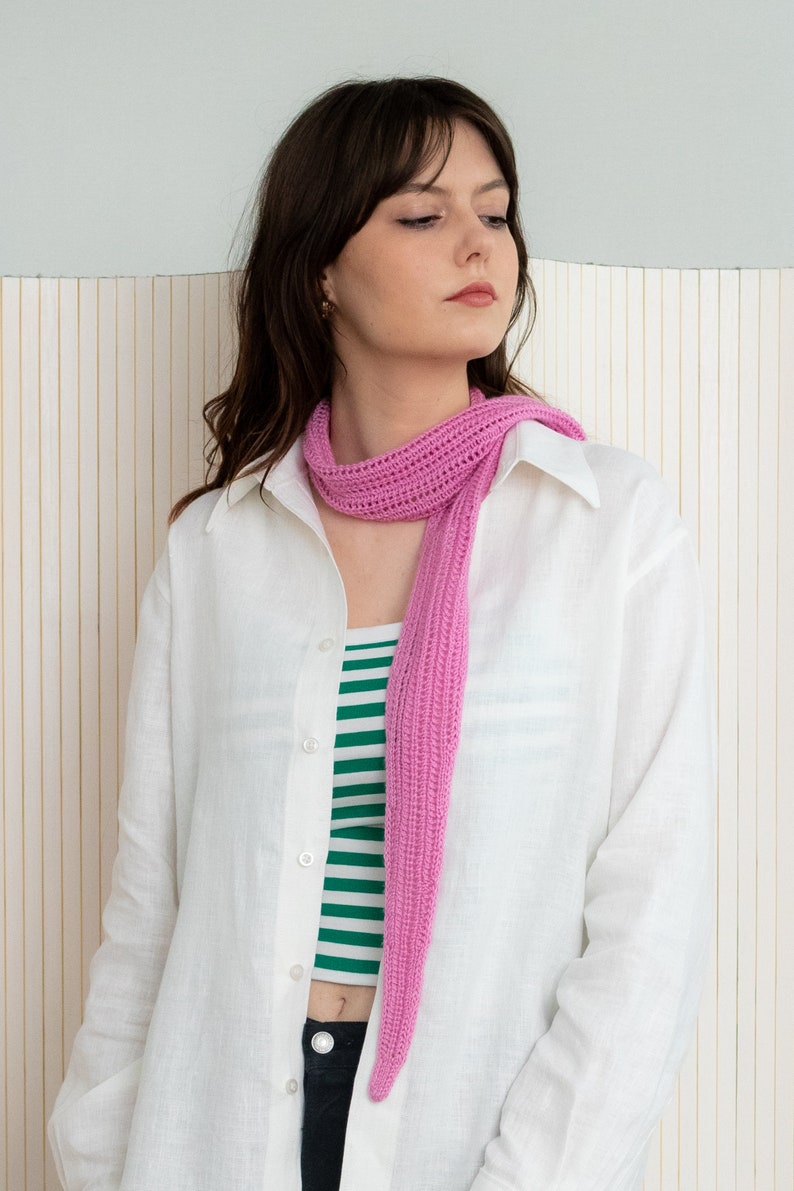 May include: A woman wearing a white linen shirt with a pink crocheted scarf. The scarf is long and hangs down to her waist.
