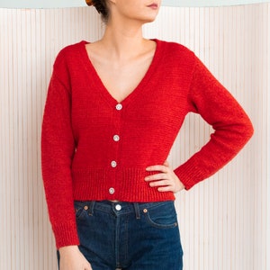 Può includere: Un cardigan rosso acceso con scollo a V e maniche lunghe. Il maglione presenta quattro bottoni scintillanti a forma di fiore sul davanti. La modella indossa jeans blu. Lo sfondo è un motivo chiaro a righe verticali.