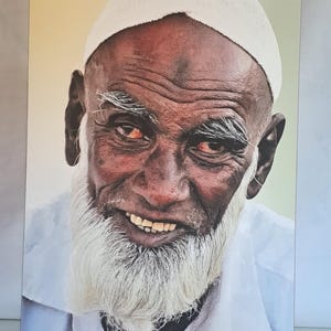 Può includere: Ritratto di un uomo anziano con la pelle scura, barba bianca e berretto bianco. L'uomo sorride, rivelando i denti. Indossa una camicia bianca. Lo sfondo è di colore giallo chiaro.