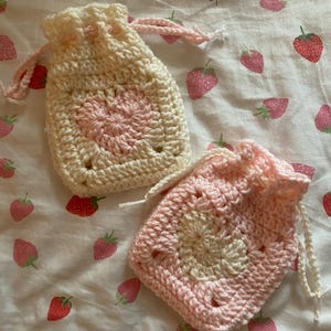 May include: Two crocheted drawstring bags with a pink and white color scheme. The bags are made with a granny square pattern and have a heart shape in the center.
