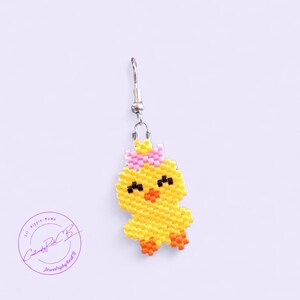 May include: A single yellow beaded chick earring with a pink bow. The chick has black eyes and an orange beak and feet. The earring is suspended from a silver-colored hook. The background is a light purple color.