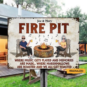 Op de afbeelding: Een gepersonaliseerd metalen bord met de tekst "Joe & Mary Fire Pit" en de zin "Where music gets played and memories are made... where marshmallows are roasted and we all get toasted". Het bord toont een cartoonafbeelding van een stel dat bij een vuurplaats zit met muzieknoten in de lucht.