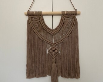 May include: A brown macrame wall hanging with a geometric design and a tassel. The hanging is made of natural fibers and has a bohemian style.