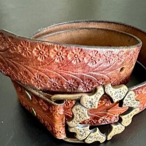 May include: A brown leather belt with a floral embossed design. The belt features a decorative buckle and is made of a rich, reddish-brown leather. The belt is a stylish accessory.