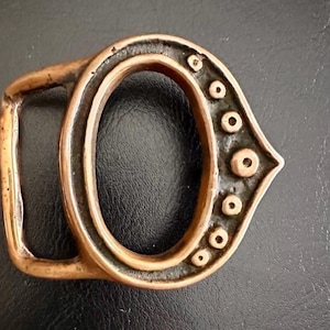 May include: A vintage-style copper-colored belt buckle. The buckle has an oval center and a rectangular loop on one side. The other side has a decorative, curved design with several small, raised circles.
