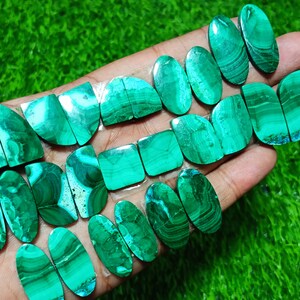 K&ouml;nnte beinhalten: Eine Sammlung von 16 gr&uuml;nen Malachit-Cabochons, einige oval, einige quadratisch und einige halbkreisf&ouml;rmig. Die Steine haben eine Vielzahl von Mustern und Texturen.
