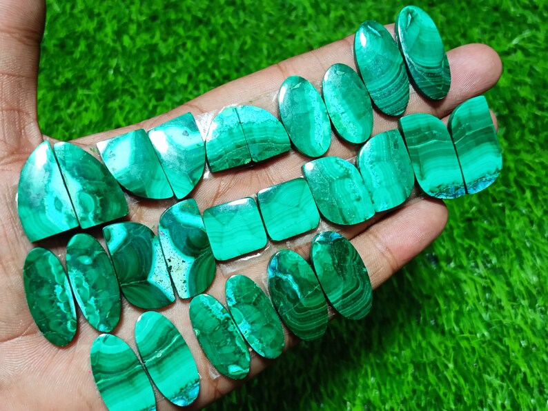 K&ouml;nnte beinhalten: Eine Sammlung von 16 gr&uuml;nen Malachit-Cabochons, einige oval, einige quadratisch und einige halbkreisf&ouml;rmig. Die Steine haben eine Vielzahl von Mustern und Texturen.