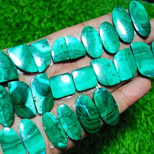 K&ouml;nnte beinhalten: Eine Sammlung von 16 gr&uuml;nen Malachit-Cabochons, einige oval, einige quadratisch und einige halbmondf&ouml;rmig. Die Steine haben eine Vielzahl von Mustern und Texturen.