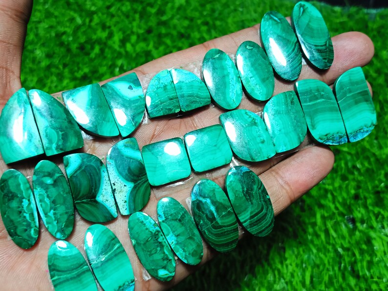 K&ouml;nnte beinhalten: Eine Sammlung von 16 gr&uuml;nen Malachit-Cabochons, einige oval, einige quadratisch und einige halbmondf&ouml;rmig. Die Steine haben eine Vielzahl von Mustern und Texturen.