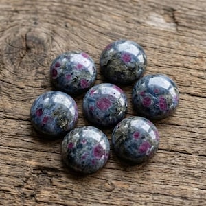 May include: Seven polished, round gemstones with a mix of dark blue-gray and red hues are arranged on a weathered wooden surface. The gemstones have a reflective, glossy finish, and the wood grain adds a natural texture.