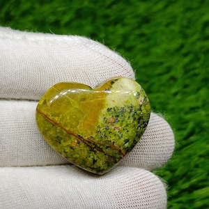 Può includere: Una gemma a forma di cuore con una superficie lucida. La pietra presenta una miscela di tonalità verde oliva, giallo e marrone, con inclusioni verde scuro e nere. Una sottile venatura marrone rossastra attraversa la pietra.