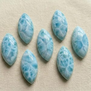May include: Seven oval-shaped larimar gemstones with a light blue and white marbled pattern. The polished stones are arranged on a beige surface, showcasing their unique textures and colors. These gemstones are ideal for jewelry making.