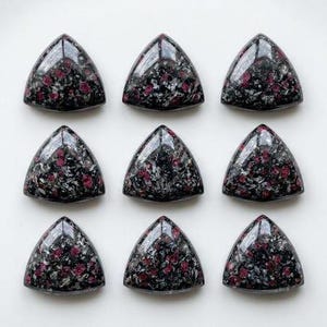 Puede incluir: Nueve cabujones triangulares pulidos dispuestos en una cuadrícula de tres por tres. Cada pieza presenta una base negra oscura con inclusiones moteadas de rojo intenso y plata metálica, creando una superficie de alto brillo y reflectante.