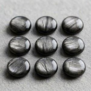 May include: Nine circular, polished black obsidian stones arranged in a three-by-three grid. Each stone features a distinct, fibrous, silver-grey chatoyant sheen that creates a shimmering, linear pattern across the smooth, rounded surface of the gems.