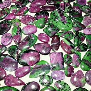 Könnte beinhalten: Eine Sammlung polierter Edelsteine in verschiedenen Formen, darunter Ovale, Tropfen und Rechtecke. Die Steine zeigen eine Mischung aus Farben, mit leuchtenden Grüntönen und tiefen Magentatönen. Die Edelsteine werden wahrscheinlich für Schmuck oder Dekorationszwecke verwendet.