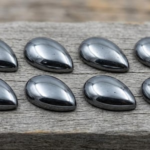 May include: Eight polished, teardrop-shaped hematite stones arranged on a weathered wooden surface. The stones have a metallic, dark gray color and a smooth, reflective surface. The teardrop shape is consistent across all stones.