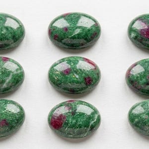 May include: Nine oval-shaped gemstones with a polished, glossy finish. The stones are a deep green color with irregular patches of red. The gemstones are arranged in a grid pattern on a white surface.