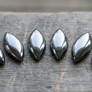 May include: Six polished, teardrop-shaped hematite stones arranged on a weathered wooden surface. The stones have a metallic, silver-gray sheen, reflecting light. The arrangement suggests a collection of decorative or craft elements.