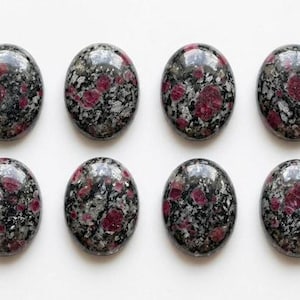 May include: Eight oval-shaped cabochons arranged in two rows of four. Each stone features a polished surface with a mottled pattern of deep magenta-red inclusions embedded within a dark gray and black speckled matrix of crystalline mineral material.