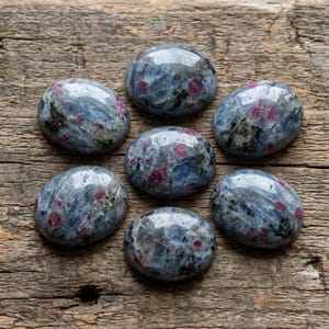 May include: Seven polished, oval-shaped gemstones with a mix of blue-grey and red hues are arranged on a weathered wooden surface. The stones have a glossy finish, and the wood grain provides a rustic backdrop.
