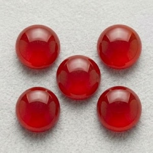Peut inclure: Cinq pierres précieuses cabochons rouge foncé, lisses et circulaires, disposées en forme de lettre H. Chaque pierre présente une surface polie et bombée qui reflète la lumière, créant un éclat brillant sur le dessus de chaque pièce.