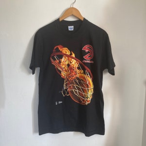 Puede incluir: Camiseta negra con un gr&aacute;fico de una pelota de baloncesto pasando por un aro en llamas. El logotipo de los Atlanta Hawks est&aacute; en el lateral del aro. El texto "Atlanta Hawks" est&aacute; debajo del logotipo.