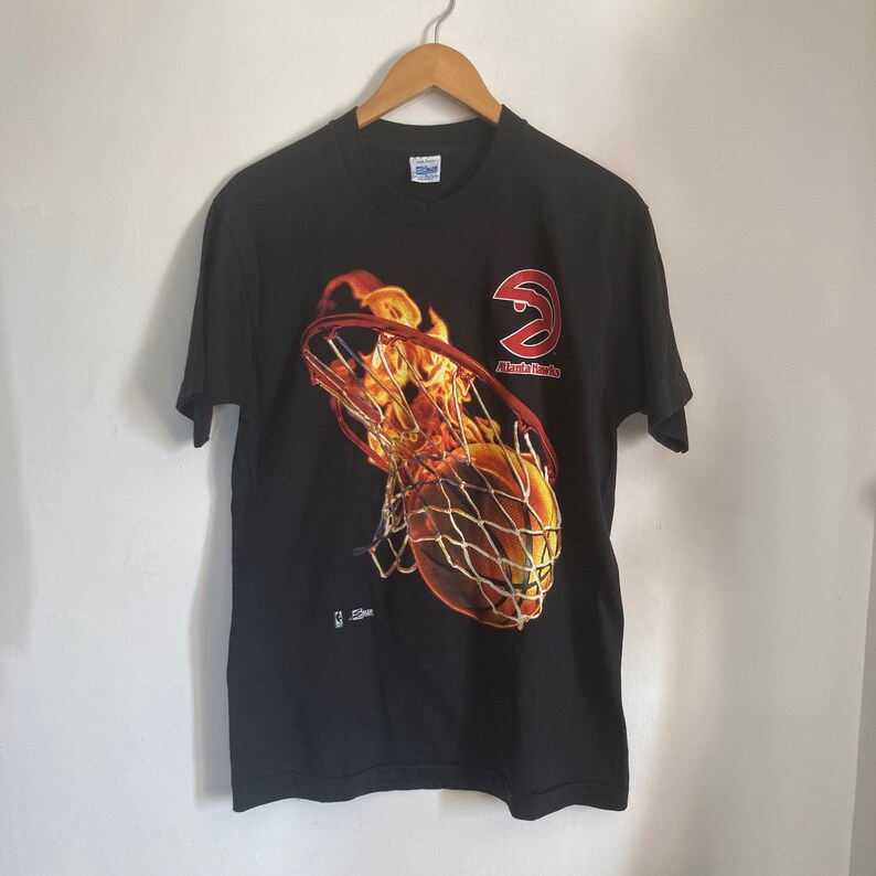 Puede incluir: Camiseta negra con un gr&aacute;fico de una pelota de baloncesto pasando por un aro en llamas. El logotipo de los Atlanta Hawks est&aacute; en el lateral del aro. El texto "Atlanta Hawks" est&aacute; debajo del logotipo.