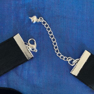 May include: A black velvet choker necklace with a silver clasp and a silver chain extender. The chain has a heart-shaped charm.