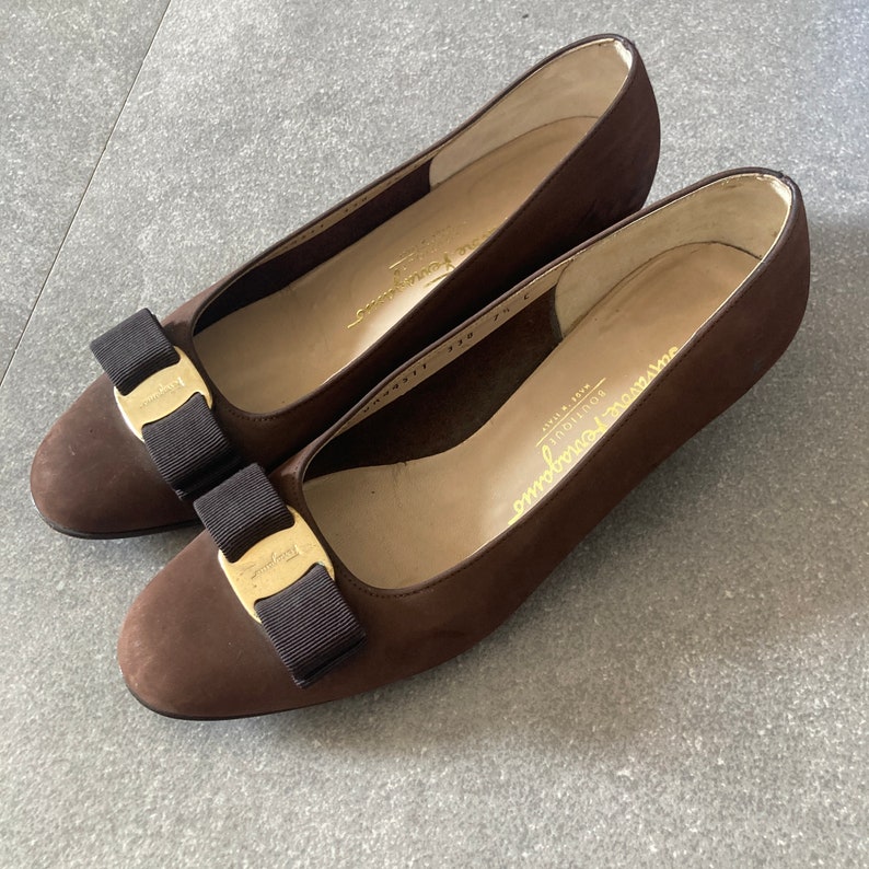 May include: A pair of brown suede heeled shoes with a gold buckle and black bow detail. The shoes have the text "Salvatore Ferragamo" and "Boutique" on the inside.