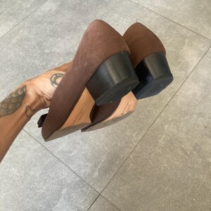 May include: A pair of brown suede heeled shoes with black heels. The shoes are worn and have some scuffs on the leather.