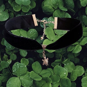 May include: A black velvet choker necklace with a silver turtle pendant. The necklace is photographed against a backdrop of green clover leaves.