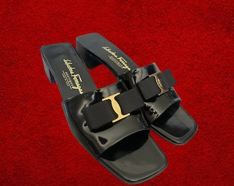 May include: Pair of black patent leather sandals with a square toe and small block heel. Each sandal features a black bow with a gold-tone buckle. The brand name "Salvatore Ferragamo" is printed on the insole.