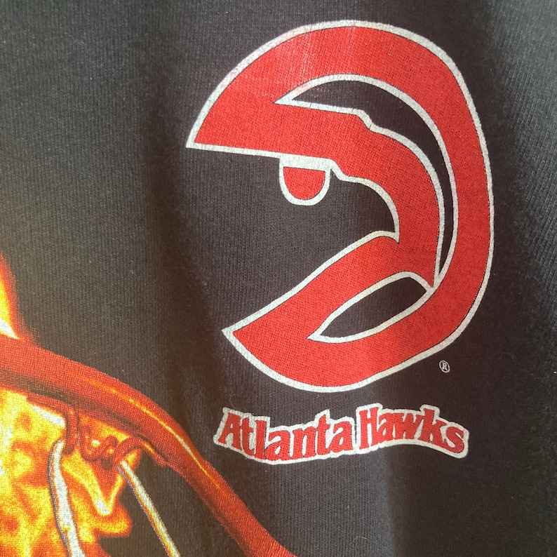 Puede incluir: Camiseta negra con un logotipo rojo de los Atlanta Hawks y el texto "Atlanta Hawks".