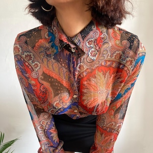 May include: A sheer, long-sleeved button-up shirt with a paisley print in shades of red, orange, blue, and brown.