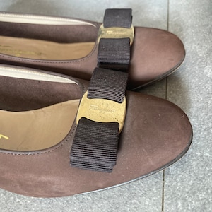 May include: A pair of brown suede ballet flats with a black grosgrain bow and a gold metal buckle that says "Salvatore Ferragamo".