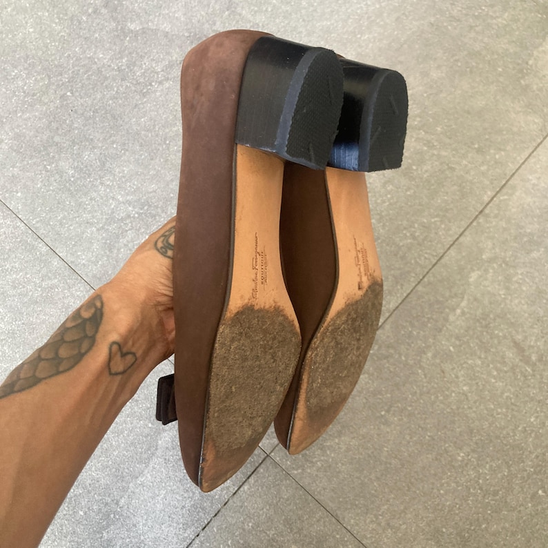 May include: A pair of brown suede heeled shoes with a black heel. The shoes have a small bow detail on the front and the brand name "Salvatore Ferragamo" is printed on the insole.