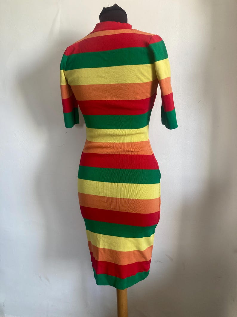 Puede incluir: Vestido ajustado a la figura con un patr&oacute;n de rayas arco&iacute;ris. El vestido tiene mangas cortas y un cuello alto. Las rayas alternan entre rojo, naranja, amarillo, verde y un amarillo m&aacute;s claro. Largo hasta la rodilla.
