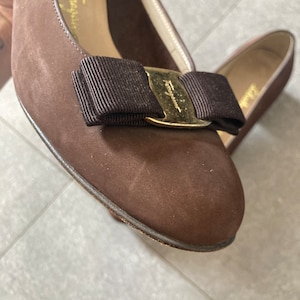 May include: A pair of brown suede shoes with a gold buckle and a black bow. The shoes have a pointed toe and a low heel.