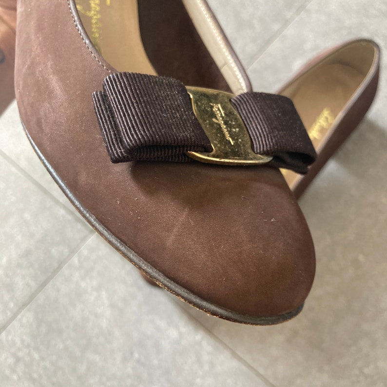 May include: A pair of brown suede shoes with a gold buckle and a black bow. The shoes have a pointed toe and a low heel.