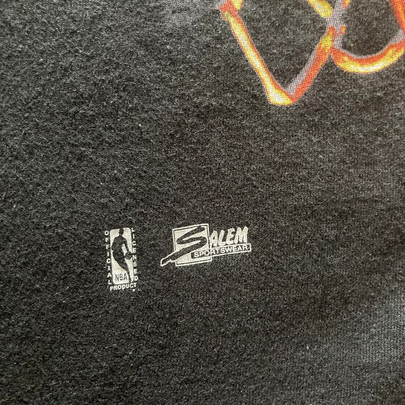 Puede incluir: Camiseta negra con un gr&aacute;fico de baloncesto y el texto "Salem Sportswear" en una etiqueta blanca. La etiqueta tambi&eacute;n incluye el logotipo de la NBA y el texto "Producto oficial con licencia de la NBA".