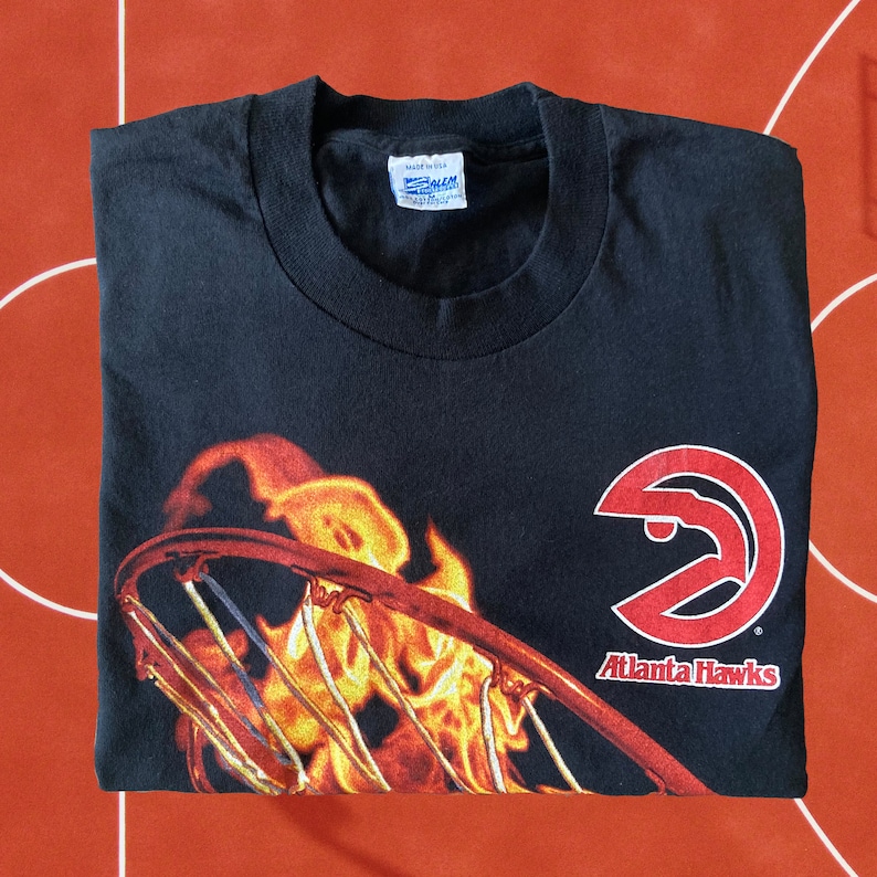 Puede incluir: Camiseta negra con un logotipo rojo de los Atlanta Hawks y un aro de baloncesto envuelto en llamas. El texto "Atlanta Hawks" est&aacute; debajo del logotipo.