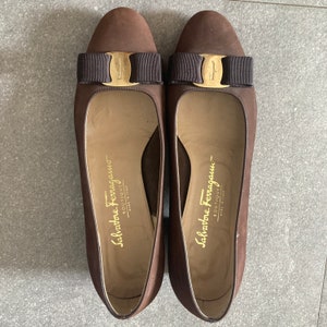 May include: A pair of brown leather heeled shoes with a gold buckle and a brown bow on the front. The shoes have the text "Salvatore Ferragamo - Boutique" on the inside.
