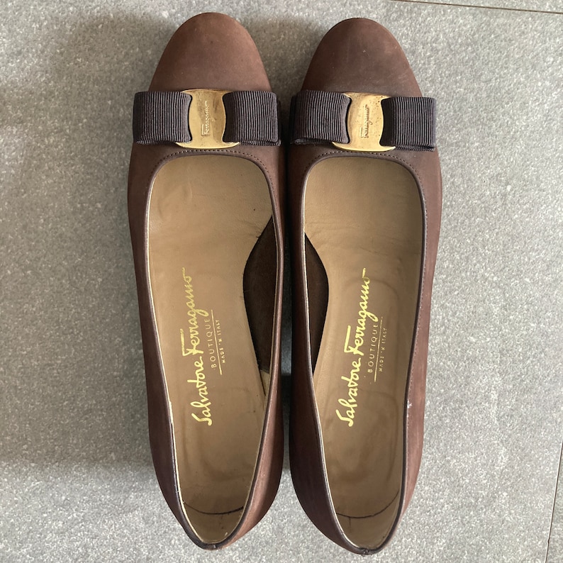 May include: A pair of brown leather heeled shoes with a gold buckle and a brown bow on the front. The shoes have the text "Salvatore Ferragamo - Boutique" on the inside.