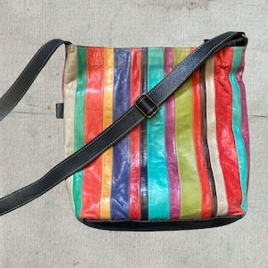 Puede incluir: Un bolso de hombro de cuero con rayas verticales que presenta una vibrante paleta de colores en rojo, naranja, amarillo, verde, turquesa, morado y crema. El bolso incluye una correa de hombro negra ajustable con hebilla metálica y base negra.