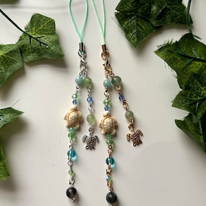 May include: Two turquoise and gold phone charms with a sea turtle design. Each charm features a white turtle charm, green and blue beads, and a dark gray bead at the bottom.