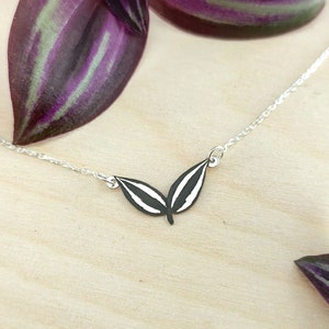 May include: A silver necklace featuring a pendant with a black and white leaf design. The necklace is displayed on a light wooden surface, with purple and green leaves in the background.