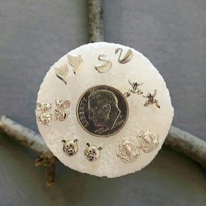 May include: A collection of small, silver-toned stud earrings in various animal shapes, including foxes, swans, frogs, pigs, and koalas. A US dime is in the center for size comparison. The earrings are displayed on a white, textured surface.