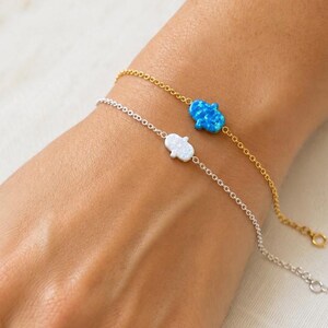 May include: Two delicate bracelets featuring Hamsa hand charms. One bracelet is gold-toned with a bright blue opal-style Hamsa charm, while the other is silver-toned with a white opal-style Hamsa charm. Both chains are thin and link-style.