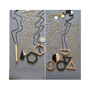 May include: A collection of geometric pendant necklaces in gold, black, and silver tones. The pendants feature shapes like triangles, hexagons, and bars. The necklaces are displayed on a gray surface.