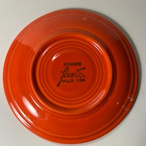 May include: A bright orange ceramic saucer with a circular recessed center. The underside features the black stamped text GENUINE fiesta H-L-O USA. The piece has a smooth, glossy finish and a raised outer rim, showing a classic round design.